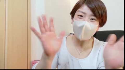 Media: Video of a woman with short brown hair, wearing a white mask and white top, waving her hands in a room with beige walls and a black chair.