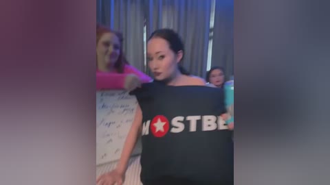 Media: A video of an Asian woman with long black hair, wearing a black \"MOSTB\" T-shirt, standing in a dimly lit room with a blurred background of people.