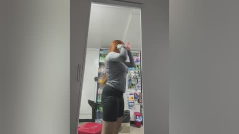 Media: Video of a red-haired woman in a tight gray long-sleeve and black shorts, playing a video game in a messy room with posters and a gaming chair.