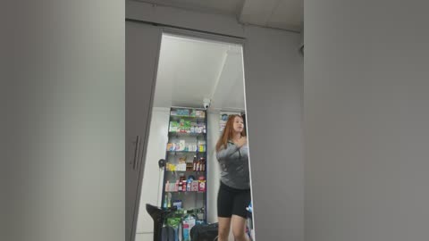 Media: Video of a woman in a grey top and black shorts, standing in a small, brightly-lit room with a white door, a refrigerator filled with colorful food items, and a plant on the floor.