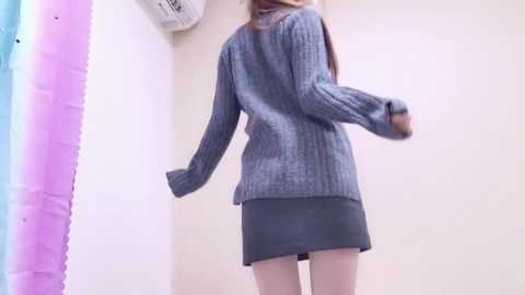 Media: A video of a young woman in a grey knit sweater and short black skirt, walking away, with a white air conditioner and pastel-colored shower curtain in the background.