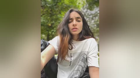 Media: Video of a young woman with long, dark hair and light skin, wearing a white T-shirt with a grey graphic and black jacket, sitting on a park bench in a lush, green park.