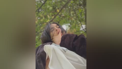 Media: Video of a young woman with long black hair, wearing a white t-shirt and dark jacket, lying on her back with eyes closed, surrounded by greenery.