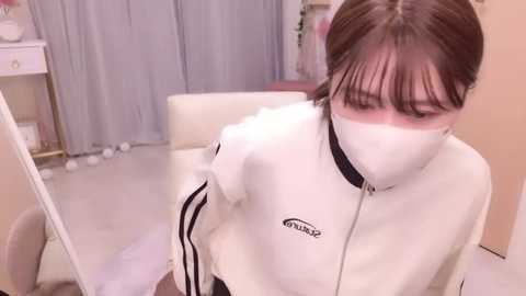 Video of an East Asian woman with fair skin, brown hair, and bangs, wearing a white tracksuit with black stripes, a face mask, and sitting on a white chair in a softly lit room with beige walls and curtains.