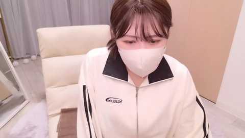 Media: Video of a young Asian woman with light skin, wearing a white face mask, white jacket, and black hair, sitting on a white leather chair, indoors with beige walls and grey curtains.