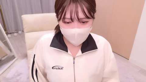 Media: Video of an Asian woman with pale skin and brown hair in a ponytail, wearing a white jacket, mask, and glasses, sitting on a white chair in a minimalist room with beige walls and a mirror.