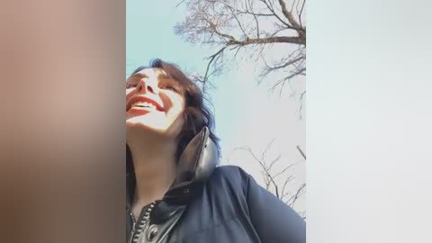 Media: Video of a smiling woman with dark hair, wearing a black jacket, outdoors with bare trees and a clear blue sky in the background.