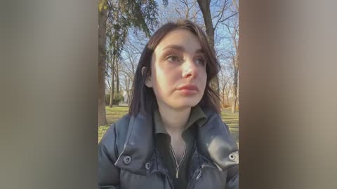 Media: Video of a young woman with fair skin, brown hair, and light makeup, wearing a black jacket and silver necklace, standing in a park with bare trees and green grass under a clear blue sky.