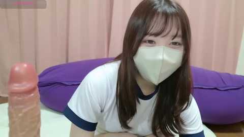 Media: Video of a young Asian woman with long brown hair and a surgical mask, wearing a white t-shirt, kneeling in a bedroom with a purple pillow and beige curtains.