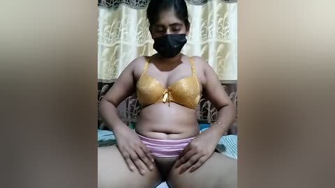 Media: A video of a South Asian woman with medium brown skin, wearing a gold lace bra and pink panties, sitting on a bed with beige curtains in the background.