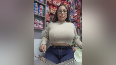 Media: Video of a Latina woman with glasses, medium skin tone, medium build, wearing a beige crop top and navy blue jeans, seated behind a counter in a convenience store.