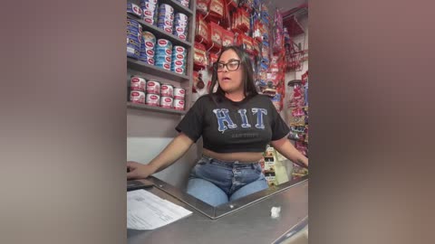 Media: Video of a young Latina woman with glasses, medium build, wearing a black \"RIT\" crop top and blue jeans, seated behind a counter in a brightly lit convenience store filled with colorful snacks and beverages.