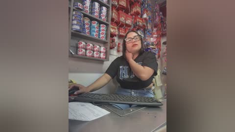 Media: Video of a middle-aged woman with glasses, wearing a black T-shirt and blue jeans, seated at a desk in a candy store, surrounded by colorful, stacked candy boxes.