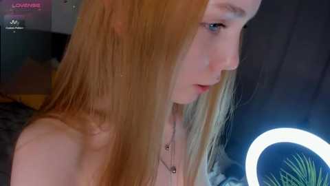 Video of a fair-skinned, red-haired young woman with long, straight hair, wearing a black top, looking down. Background shows a dimly lit room with a bright ring light and a plant.