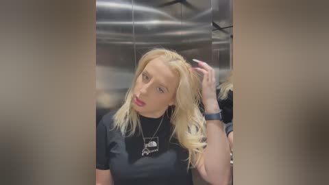 Media: Video of a blonde woman with fair skin and pink lipstick, wearing a black top and silver necklace, in a mirrored elevator. She touches her hair while gazing at her reflection.