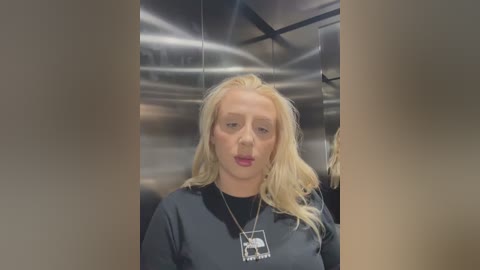 Video of a blonde woman with pale skin, wearing a black hoodie and a silver pendant necklace, standing in an industrial, metallic elevator with reflective surfaces.