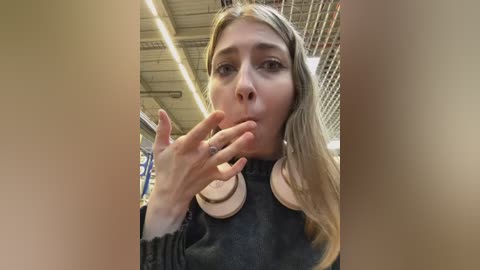 Media: A video of a blonde woman in a black sweater, holding a cigarette to her mouth, taken in a dimly lit warehouse with industrial ceiling and metal beams.