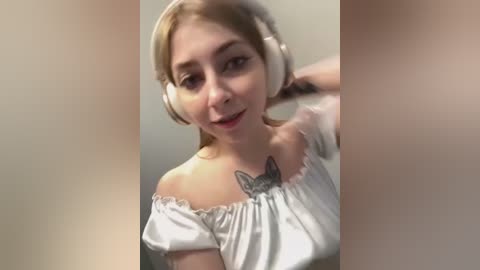 Media: Video of a young woman with light skin, blonde hair, and a butterfly tattoo on her chest. She wears a white off-shoulder blouse and large headphones. The background is blurred.