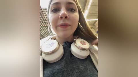 Media: Video of a young Caucasian woman with light skin and long brown hair, wearing gray sweater, large white over-ear headphones, and a neutral expression. Background shows a beige ceiling with metal grid.