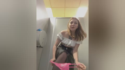 Media: Video of a young woman in a public restroom, removing pink panties, wearing a white off-shoulder blouse and black skirt.