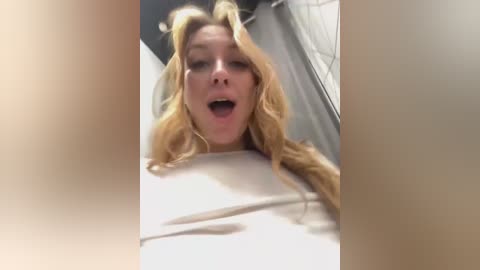 Media: A blurry video of a young, blonde woman with wavy hair, wearing a white top, looking surprised or startled, taken from a low angle. Background includes a shower curtain and a bathroom sink.