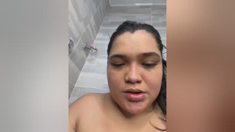 Media: Video of a young Latina woman with medium skin tone, wet hair, and closed eyes, standing in a shower with light gray tiled walls, looking upwards.