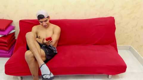 Media: Video of a shirtless young man with medium skin tone, wearing a backwards white baseball cap, black shorts, and grey sandals, sitting on a red couch, holding a red smartphone, with a stack of red and purple clothes in the background.