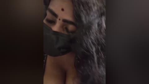 Media: Video of a close-up of a South Asian woman with medium brown skin and long, wavy black hair, partially covered by a black mask. Her eyes are closed, and she has a red bindi on her forehead. The background is dark and blurry.