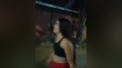 Media: A blurry video of a woman with long, dark hair wearing a black sports bra and red shorts, walking in a dimly lit, rustic outdoor area with wooden structures and a red brick wall.