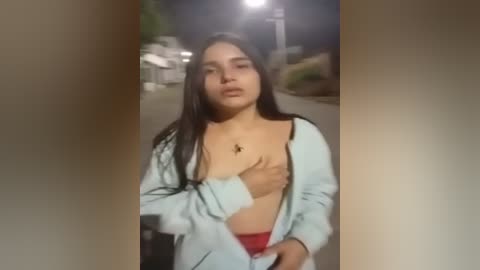 Media: Video of a young Latina woman with long dark hair, wearing a light blue hoodie, exposing her breasts, standing in a dimly lit street at night.