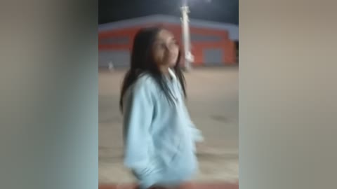 Media: A blurred video of a young person with long black hair, wearing a light blue jacket, standing in front of a red and orange building at night.