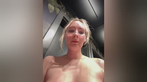 A video of a topless blonde woman with glasses, fair skin, and medium-sized breasts, standing in a modern bathroom with a green plant on the left.