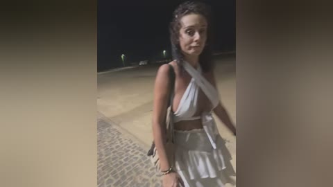 Media: Video of a woman with medium skin tone, long dark hair, wearing a white halter top and pleated skirt, standing on a cobblestone path at night.