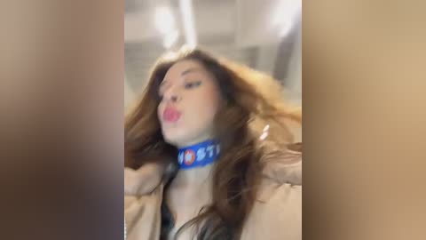 Media: A blurry video shows a woman with long brown hair, wearing a blue collar with white text, red lipstick, and a beige jacket, in a dimly lit room.