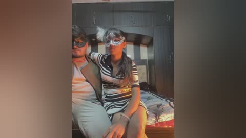 Media: Video of a man and woman in a bedroom. The man wears a striped shirt, and the woman has a striped shirt and a face mask, both sitting on a bed.