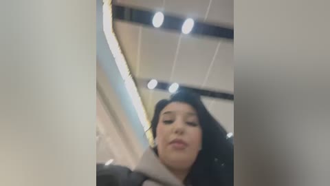 Media: Video of a young woman with long black hair, light skin, and closed eyes, looking up. She wears a dark jacket. Background shows a modern, well-lit room with a curved mirror, ceiling lights, and white walls.