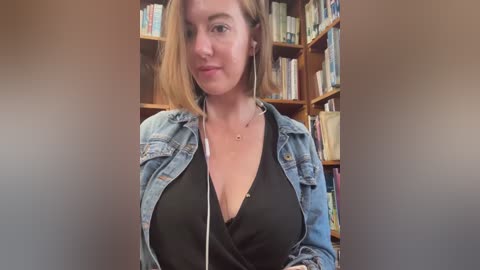 Media: A video of a fair-skinned woman with blonde hair, wearing a black top and denim jacket, standing in a book-filled room. She has a neutral expression.