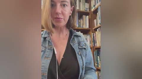 Media: Video of a woman with shoulder-length, light brown hair, wearing a black top and light blue denim jacket, standing in a room filled with bookshelves.