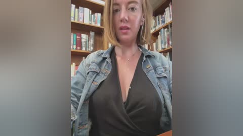 Media: Video of a blonde woman with a light complexion, wearing a black dress under a denim jacket, standing in front of a bookshelf filled with books.