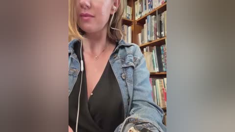 Media: Video of a fair-skinned woman with blonde hair, wearing a black V-neck top and denim jacket, listening to music with white earbuds, in a library with wooden bookshelves filled with books.