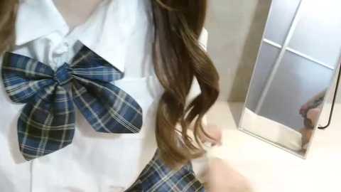 Media: Video of a young person with long, wavy brown hair wearing a white shirt with a large, plaid bow tie, adjusting their hair in a mirror.