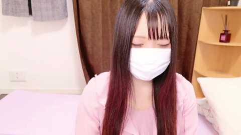 Video of an Asian woman with long black hair, wearing a white surgical mask, sitting on a bed in a room with light brown curtains and wooden shelves holding a red bottle.