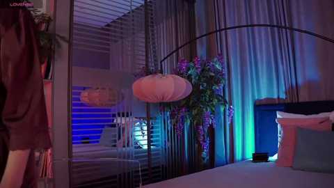 Media: Video of a modern bedroom with a bed covered in white linens, purple and blue lighting, a hanging paper lantern, and a potted plant near a window.