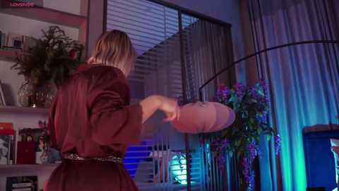 Media: Video of a woman in a maroon robe throwing a stuffed animal against a window with blinds. Background includes a bookshelf, plants, and a TV, with blue and purple lighting.