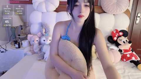 Media: Video of an Asian woman with long black hair, wearing a blue bikini, hugging a large stuffed animal on a bed with white pillows and stuffed Minnie Mouse and Donald Duck toys.