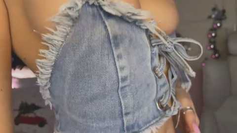 Media: Video of a woman's upper torso wearing a distressed denim halter top with frayed edges and a tied front. Her skin is light, and she has a silver bracelet on her wrist. Background shows a blurred, indoor setting with a pink and purple beaded necklace hanging.