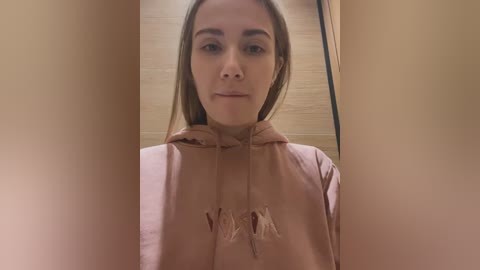 Media: Video of a young woman with light skin, straight brown hair, and brown eyes, wearing a beige hoodie with tassel accents. Background features a light wooden wall. She has a neutral expression.