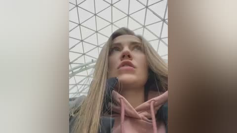 Video of a young woman with long, straight blonde hair, wearing a pink hoodie, gazing upward. The background features a geometric, grid-like glass roof. Her expression is contemplative.