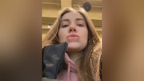 Media: Video of a young woman with long, light brown hair, fair skin, and a neutral expression. She is wearing a pink hoodie and holding a black pouch. The background is blurry, showing an indoor setting with ceiling beams.