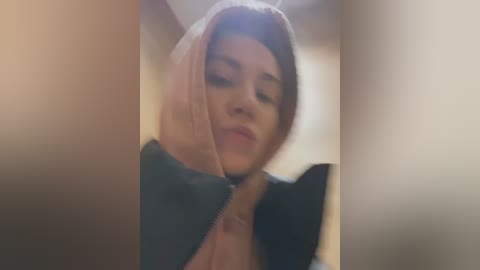 Media: A blurry, low-resolution video of a person wearing a hooded jacket with a pink lining, partially obscured by soft-focus, beige-colored objects.